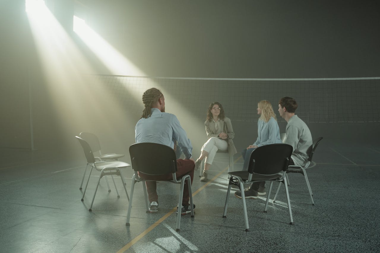A group of diverse adults in a support meeting in a sunny indoor gym setting.